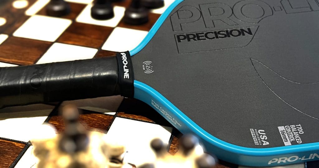 Close-up of Pro-Line Precision 16mm pickleball paddle resting on a chessboard, highlighting its textured face, ergonomic grip, and USA Pickleball approved control-focused design
