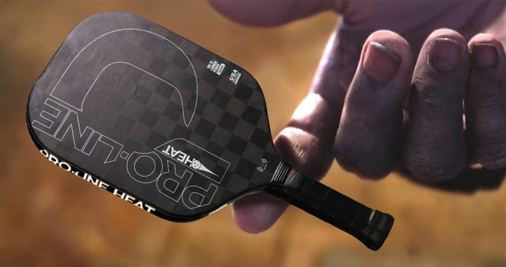 Close-up of a large hand holding a Pro-Line Heat pickleball paddle with a carbon fiber face, edgeless design, and bold branding, styled dramatically like a powerful weapon being handed off.