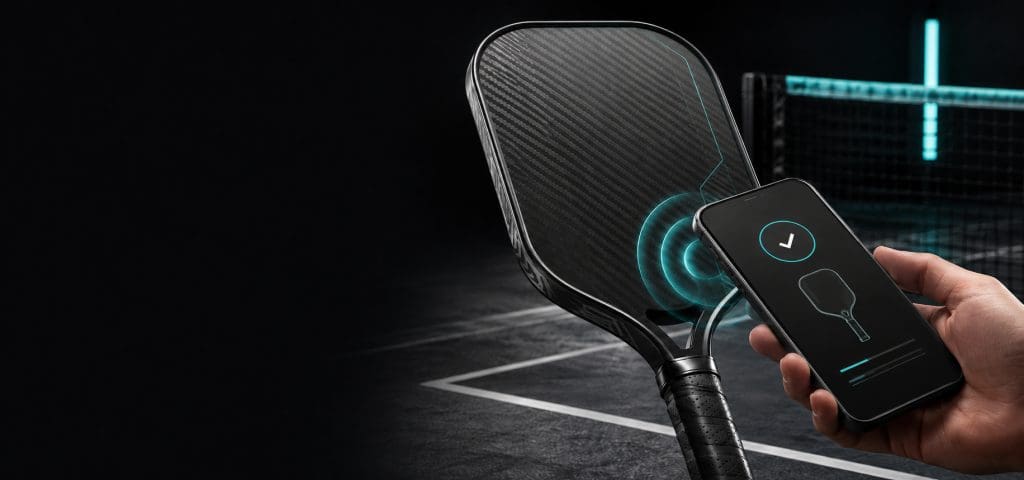 Black carbon-fiber pickleball paddle connecting to a smartphone through turquoise NFC registration technology on a dark court.